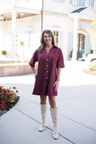 Woman posing outdoors in a rich burgundy faux-suede shirt dress featuring short elbow-length cuffed sleeves, a notched collar, and a full-length front placket with decorative gold crest buttons. The shift-style mini dress is styled with tall beige suede block-heel boots, layered gold link chain necklaces with an open-oval ring pendant, and gold statement jewelry against an architectural courtyard backdrop.