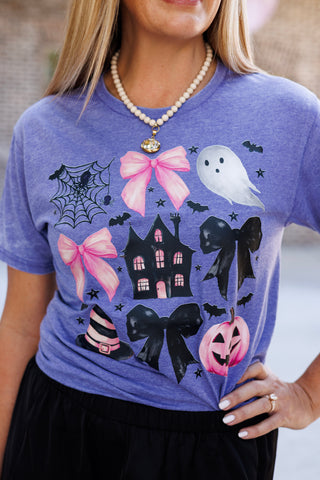 Macro close-up shot focusing on the chest print of the soft heathered purple t-shirt. Shows the crisp contrast of the girly pink bows, black haunted house with glowing pink windows, white ghost, spiderweb with a spider, black ribbon bows, striped witch hat, pink carved pumpkin, and scattering of tiny stars and bats.