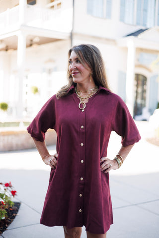 Three-quarter portrait view of a woman standing with hands on her hips in a deep wine-red faux-suede tunic dress. Highlights the soft, matte suede texture, relaxed drop shoulders, cuffed sleeves, and metallic gold button accents running down the center front.