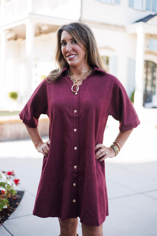Close-up portrait view of a smiling woman showcasing the notched V-neckline and open collar of a burgundy faux-suede button-front dress. The rich fabric texture is complemented by stacked gold chain necklaces, an oval pendant, and gold drop earrings.