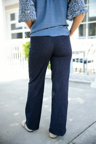 Back view of the high-waisted dark wash flare jeans, showcasing the fitted hips, back yoke construction, and classic patch pockets. The image highlights the smooth, wide-leg flare silhouette extending to a clean hemline over neutral pointed-toe flats on an outdoor concrete walkway.