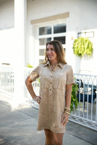 A close-up view of a woman wearing a light tan faux suede short-sleeve mini dress. The dress features a spread collar, a partial button-down placket with bronze snap buttons, and structured chest flap pockets. Styled with gold chunky jewelry including stacked bangles and a crescent-pendant chain necklace.