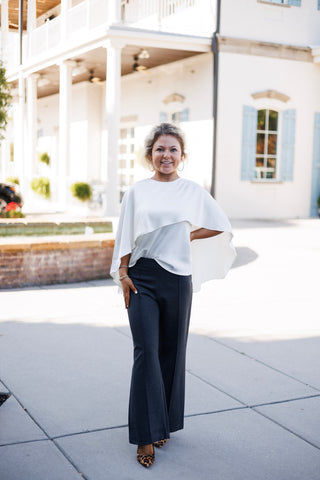 Woman posing outdoors in an elegant ivory satin-blend cape top featuring a asymmetrical draped capelet overlay across the chest and open flutter sleeves. The top is paired with high-waisted charcoal grey wide-leg trousers with center seam details, styled with leopard-print pointed-toe pumps and gold hoop earrings against a bright architectural background