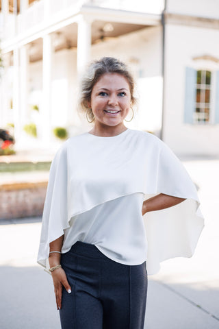 Three-quarter portrait of a smiling woman wearing an ivory capelet blouse with a high crew neckline and a fluid, sweeping cape sleeve silhouette. Posed with one hand on her hip, highlighting the clean blouson drape at the waistband of dark grey trousers.