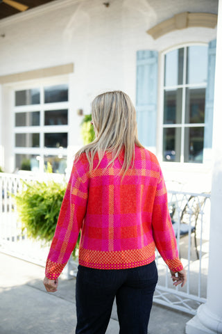 Rear view of a woman showcasing the back of a bright pink and orange plaid zip-up knit jacket. Highlights the structured grid pattern across the back and long sleeves, finished with a geometric patterned trim along the cuffs and hemline, worn with dark-wash jeans.