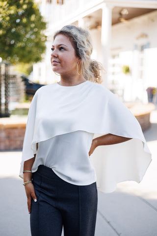 Close-up angled portrait showcasing the soft satin texture and cascading chest overlay of an ivory short-sleeve cape top. Accessorized with large gold hoop earrings and delicate stacked bracelets.