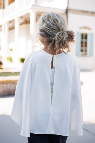 Back view of the ivory cape top highlighting the dramatic open slit design across the rear capelet and a single keyhole button closure at the neck. The model stands with her back turned, showing the elegant fluid drape of the split cape back over dark grey trousers.
