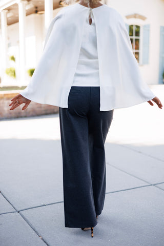 Back view of the outfit showcasing the open-back slit design of the ivory cape top with a keyhole button closure at the nape. The model holds her arms out to reveal the flowing split cape sleeves and the sleek back paneling of the charcoal wide-leg trousers.