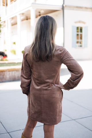 Back view of a woman wearing a taupe brown faux-suede tunic dress showcasing the clean, tailored back seam construction and long sleeves. One hand rests on her waist, highlighting the comfortable shift drape and knee-skirting hem.