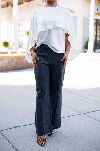 Three-quarter front view focusing on the fluid drape of an ivory capelet blouse and tailored dark grey wide-leg trousers. Highlights the clean flat waistband, front crease seams of the pants, and leopard print pumps peeking beneath the hem.