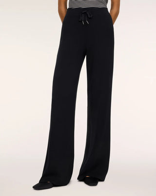 Front view of a model wearing the Very Black AirEssentials wide-leg pant paired with pointed-toe black flats and a tucked-in black-and-white striped top. The high-waisted lounge trousers feature a smooth, flat elastic waistband with an adjustable drawstring tie accented by silver aglets, flowing into an elongated, drapey wide-leg cut against a seamless white studio backdrop.   