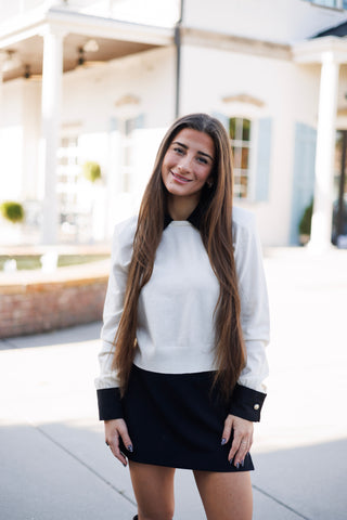 Woman smiling outdoors on a paved courtyard, wearing a cream fine-knit sweater top featuring a preppy layered look illusion with a crisp black point collar and matching extended black buttoned wrist cuffs. Styled with a black mini skirt and long straight brown hair worn loose in front of a white building.