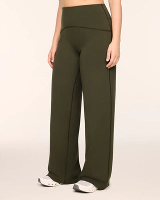 Woman modeling high-waisted wide-leg activewear pants in a deep monstera olive green. The design features a wide, smoothing waistband and prominent dark contrasting seams that curve down the front of the legs to elongate the silhouette. The fluid, relaxed-fit fabric falls cleanly to the floor, styled with chunky white athletic sneakers against a solid light studio background for a sleek, modern athleisure aesthetic.