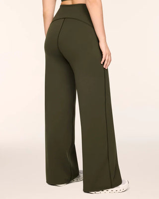Back view of the monstera olive green wide-leg pants, highlighting the supportive high-rise fit and a flattering curved back yoke seam engineered to naturally contour the glutes. The distinct dark topstitching detail continues straight down the back of each leg, emphasizing the comfortable, structured drape of the wide-leg opening as it rests effortlessly over white running shoes.