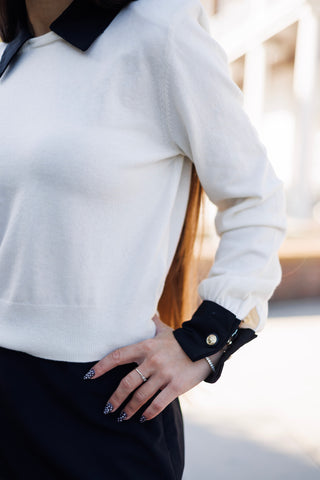 A detailed close-up shot focusing on the waist and sleeve cuff of the cream sweater, highlighting the soft fine-gauge knit fabric, ribbed bottom band, and the black woven cuff accent detailed with a gold engraved button and cuff slit.