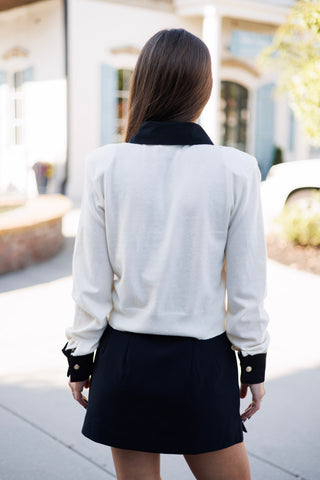 Back view from the waist up showcasing the clean, straight fit of the cream fine-knit pullover sweater. Details include the fold-over black collar extending across the back neck seam, structured shoulder seams, and wide black cuff trims with gold buttons.
