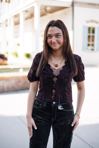 Woman smiling outdoors wearing a dark plum purple short-sleeve lace blouse with an intricate paisley pattern. Features a scalloped V-neckline, short puff sleeves, and a front keyhole button-loop closure worn layered over a lace camisole. Styled with high-waisted black corduroy pants with contrast white stitching.