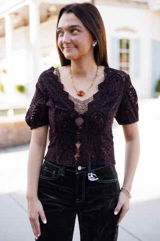 Three-quarter portrait of a woman posing in a deep plum purple short-sleeve paisley lace top. Showcases the scalloped neckline, sheer textured lace overlay, central keyhole cutout button details, paired with a gold link chain necklace with a amber pendant stone and dark velvet corduroy pants.