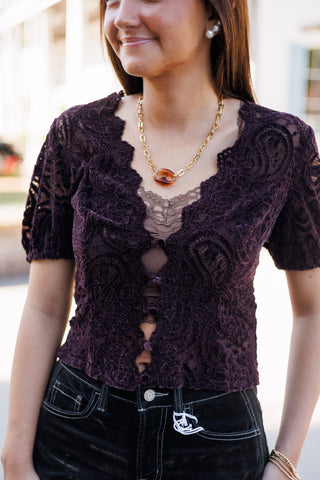 Close-up shot of the dark plum purple short-sleeve lace blouse emphasizing the intricate paisley lace weave, scalloped edge V-neck, and front fabric-covered button closures over a brown lace camisole. Styled with a gold statement chain necklace and pearl earrings.