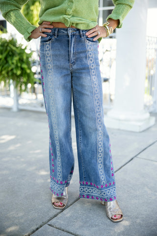 Lower body shot focusing on medium-wash wide-leg denim jeans featuring intricate white eyelet-style embroidery and vibrant magenta floral stitching running down the front seams and framing the lower hemline. The pants feature a mid-to-high waist with front scoop pockets and are styled with a light lime green cropped cardigan and open-toe metallic gold block-heel sandals on an outdoor concrete walkway.