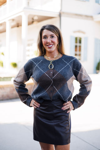 Three-quarter front view of a smiling woman wearing a long-sleeve argyle button-down cardigan in grey, brown, and beige tones tucked into a high-waisted black leather mini skirt. Accessorized with stacked gold chain necklaces, a open-oval ring pendant, and statement rings.