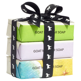 A stacked set of three individually wrapped artisan goat milk soap bars tied neatly with a black grosgrain ribbon styled into a bow on top. The ribbon features a repeating white silhouette pattern of a standing goat. The top soap bar is wrapped in lime green paper with bold black text reading "GOAT MILK BAR SOAP". The middle bar is wrapped in light lavender paper with matching text. The bottom bar features a beach-themed wrapper with blue water, sandy shores, and a red starfish image. The stack is isolated 