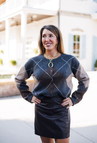 Close-up front view of a woman in a charcoal grey and brown argyle button-up knit sweater paired with a black faux-leather skirt. Styled with layered gold jewelry including a prominent oval pendant necklace, showing off the crewneck button closure and cozy knit texture.