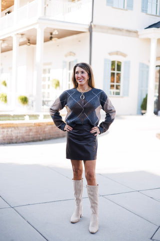 Woman posing outdoors in a charcoal gray, brown, and tan argyle knit button-front cardigan sweater tucked into a black faux-leather mini skirt. She stands with hands on her hips wearing knee-high beige suede boots, styled with layered gold necklaces with a bold oval pendant and gold dangling earrings against a bright architectural background.