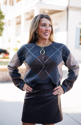 Woman posing outdoors in an argyle knit button-front cardigan sweater styled in charcoal grey, dark brown, and tan tones, tucked into a structured brown faux-leather mini skirt. She stands smiling with hands on her hips, wearing layered gold chain necklaces with a bold oval ring pendant and gold drop earrings against a bright architectural backdrop.