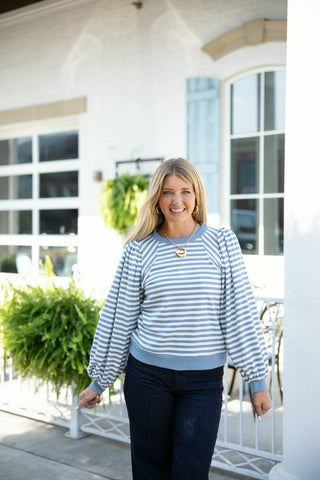 A blonde woman smiling outdoors wearing a dusty blue and white horizontal striped sweatshirt top with dramatic, voluminous raglan puff sleeves. The pullover feature solid light blue ribbed trim around the crew neckline, hem, and fitted sleeve cuffs. Styled casually with dark denim jeans, gold hoop earrings, and a thick gold chain necklace with a round locket pendant in front of a white building patio.