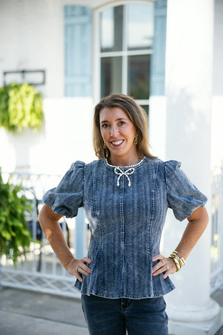 Portrait shot of a woman wearing an acid wash blue corduroy crop top featuring short puff sleeves, structured vertical seam lines, and a flared peplum waist hem. The round crew neckline is detailed with a decorative pearl bead embellishment and an attached pearl bow motif at the center chest. Accessorized with gold drop earrings and stacked gold bangles against an outdoor patio setting.