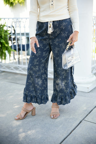 A close-up shot highlighting the washed denim texture and subtle floral print of cropped wide-leg pants. Detail view showcases the cream tasseled drawstring waist, frayed side-seam ruffles, tiered ruffle hem with raw edges, tan braided dual-strap heeled sandals, and a clear vinyl handbag with a chunky gold chain handle.
