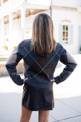 Back view of a woman wearing a charcoal grey and brown argyle knit cardigan sweater tucked into a black faux-leather mini skirt. She stands with hands on her waist, showcasing the geometric diamond pattern across the back and long sleeves.