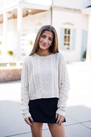 Front view of a woman wearing a relaxed-fit cream cable-knit crop sweater paired with a dark black skirt. Highlights the cozy, textured knit detailing across the chest and sleeves, ribbed crew neckline, and cropped boxy silhouette.