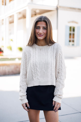 Woman smiling outdoors in a cream-colored, thick cable-knit cropped sweater layered over a black mini skirt. The oversized crewneck sweater features intricate mixed cable braid patterns, dropped shoulders, long sleeves, and ribbed trim at the hem and cuffs. Styled in front of a modern white building courtyard.