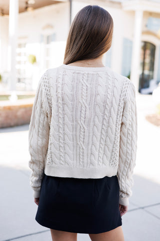Back view of the cream cable-knit sweater displaying the continuous cable weave pattern across the shoulders and back. The cropped straight ribbed hem rests above a black mini skirt, demonstrating the cozy knit depth and relaxed rear fit.