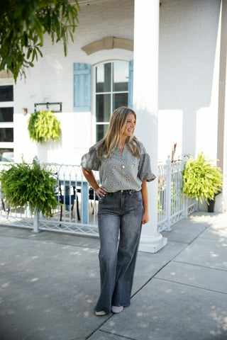 A blonde woman posing outdoors in high-waisted, grey washed wide-leg jeans paired with a sage green short-sleeved embroidered blouse. She is standing on a sunlit concrete patio with one hand on her hip, styled with a long pendant necklace and light beige pointed-toe flats in front of a white building with blue shutters and lush hanging ferns.