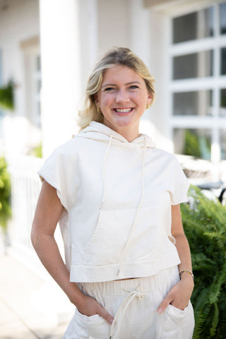 Woman smiling outdoors while modeling the French Vanilla Set's top, a solid warm cream short-sleeve hoodie. The image highlights the top's slightly cropped, boxy fit with cap sleeves, adjustable neckline drawstrings, and a spacious kangaroo front pocket, creating a cozy and structured transitional autumn look.