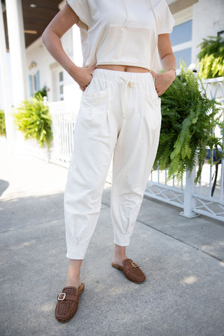 Front view of the French Vanilla Set jogger pants in a warm cream hue. The relaxed-fit bottoms showcase a comfortable high-waisted elastic drawstring waistband, deep side slash pockets, and uniquely gathered, tapered ankles. The outfit is styled with brown woven leather mules on an outdoor sidewalk.