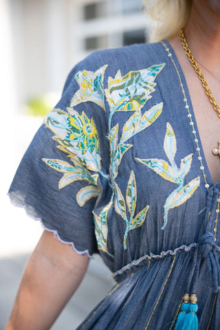 A close-up view of the Embroidered Chambray Tiered Maxi Dress highlighting the textured, artisanal details of the garment. The image focuses on the vibrant white, yellow, and green leafy applique embroidery on the shoulder and bodice. The textured blue fabric is accented with delicate gold vertical stitching, scalloped sleeve hems with white embroidered edges, and a V-neckline finished with a braided string tie featuring gold beads and bright blue tassels.