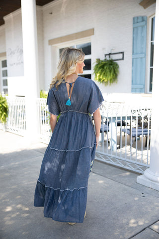 Back view of the Embroidered Chambray Tiered Maxi Dress showcasing a V-shaped back neckline secured across the top with a matching blue tassel tie. The image highlights the relaxed, flowing silhouette of the tiered maxi skirt, featuring subtle frayed ruffle details along the horizontal seams, and airy flutter sleeves. The model is standing on a bright outdoor patio, highlighting the dress's lightweight, breathable drape.
