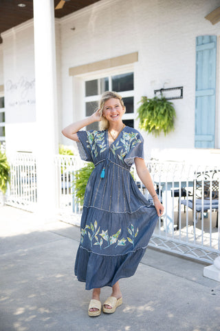 Woman smiling and posing in the Embroidered Chambray Tiered Maxi Dress. The bohemian-inspired dress features a deep V-neckline, flutter sleeves with scalloped edges, and an empire waist cinched with a vibrant blue tassel tie. The long, tiered skirt and upper bodice showcase intricate, botanical applique embroidery in shades of white, green, and yellow. Styled for a breezy, summer aesthetic with woven raffia platform sandals and layered gold jewelry, set against the backdrop of a white brick building with li