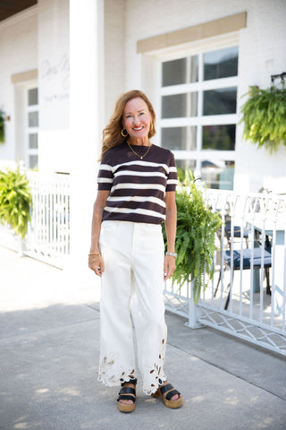 Woman smiling and posing in a chic, casual outfit featuring a dark brown short-sleeve knit sweater with alternating thick white and thin tan horizontal stripes. The top is paired with high-waisted, white wide-leg pants that showcase striking, large floral cutout eyelet embroidery around the bottom hem. Styled with black strappy platform sandals, gold hoop earrings, and a delicate gold pendant necklace, standing outdoors on a sunlit patio in front of a white brick building with pillars.