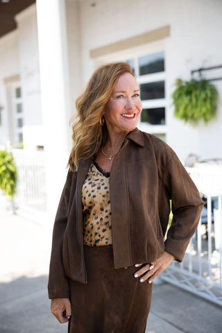 A portrait view of a woman smiling brightly with her hand on her hip, showcasing the relaxed, slightly cropped fit of the dark brown faux-suede jacket. Worn effortlessly open, the jacket features a classic pointed collar, clean front seaming, and a smooth, unembellished drape that perfectly frames the leopard print lace-trim camisole underneath. The matching faux-suede skirt and layered gold jewelry complete this cohesive, elevated transitional seasonal outfit.