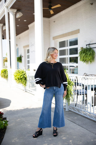 Woman smiling and looking to the side, posing in the High-Waisted Wide-Leg Cropped Jeans. This medium-wash denim features a flattering high rise, a relaxed wide-leg silhouette, and a trendy raw frayed hem. Styled for a casual, elevated everyday look with a black long-sleeve pullover featuring white varsity stripes on the sleeves, and black strappy platform wedge sandals. The scene is set outdoors on a sunlit patio in front of a modern white brick building with tall columns.