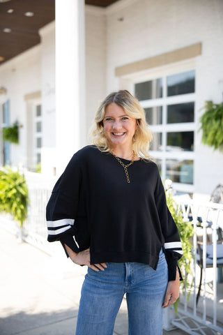 Woman smiling with her hand on her hip, posing in the Black Varsity Stripe Pullover. This casual-chic black long-sleeve top features a classic crew neckline, a relaxed drop-shoulder fit, and sporty double white stripes wrapping around the forearms. Styled for an everyday elevated look with high-waisted wide-leg blue denim jeans and a gold link Y-necklace, set outdoors on a sunlit patio in front of a white brick building.