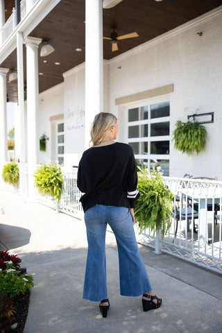 Back view of the Black Varsity Stripe Pullover and wide-leg cropped jeans outfit. The image displays the clean, unembellished back of the relaxed-fit black sweater, emphasizing the slight drape of the hemline and the sporty white sleeve stripes visible from behind. The look is completed with black strapped platform wedge sandals.