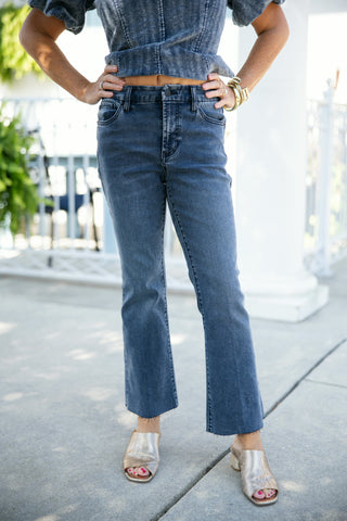Front view of a woman posing with hands on hips in a matching washed blue denim set, featuring high-waisted cropped flare jeans with raw-edge hems and classic front pockets. The jeans are paired with a matching acid-wash denim top with puff sleeves, styled with metallic gold block-heel mules and stacked gold bangles on an outdoor patio setting.