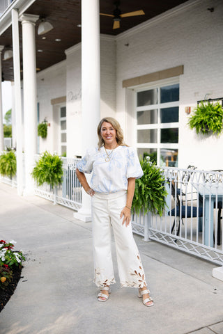 Woman posing outdoors in the Seaside Stitchery Blouse, an elegant white woven top adorned with oversized, stylized coastal blue floral embroidery outlined in a subtle cream-toned accent thread. The blouse features a classic crew neckline and voluminous short puff sleeves with soft gathering at the shoulders. It is styled for a high-end resort aesthetic, paired with cream-colored wide-leg trousers that feature intricate laser-cut floral embroidery.
