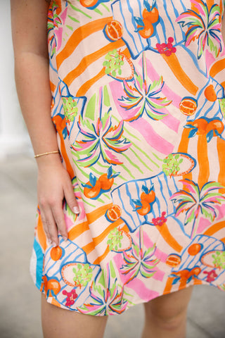 A detailed close-up shot of the high-quality woven fabric of the Citrus Cabana Mini Dress, focusing on the heavily saturated "Tangerine Mix" pattern. The print features artistic swaying palm trees, refreshing citrus slices, and colorful pink and orange diagonal stripes. The smooth texture of the fully lined fabric is visible as the model rests her hand on the side, showcasing the clean, premium finish of the textile.